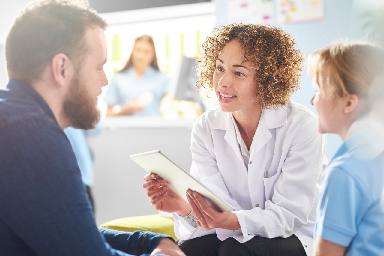 A female pharmacist sits with a father and sick daughter in the pharmacist consultation area and discusses his prescription and choice of medication viewing the details on a digital tablet, in the background a senior woman and is at the counter being served by a female pharmacy assistant