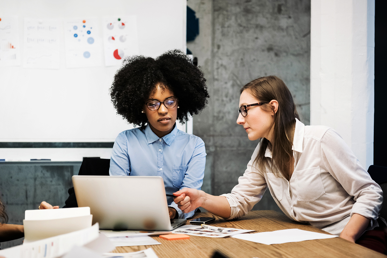 A black and a caucasian young woman are sitting in a business meeting in a modern office.They discuss something over papers and a laptop while one of them is pointing at the computer screen. Both are wearing glasses and pie charts are visible in the background.