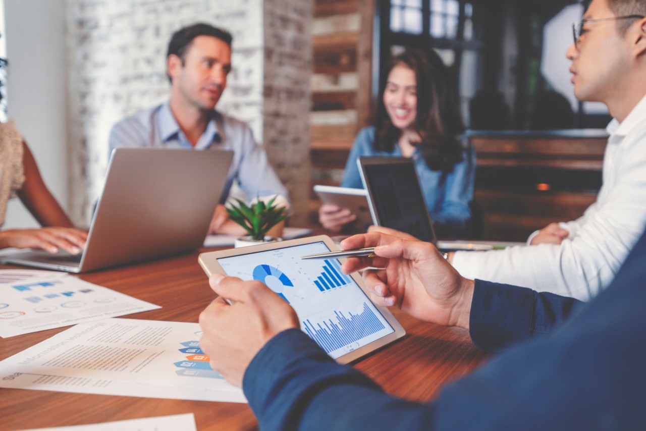 Group of people meeting with technology. There are marketing, financial and strategy documents on the wooden board room table and on the computers and digital tablets. Multi ethnic group including Caucasian and African American.